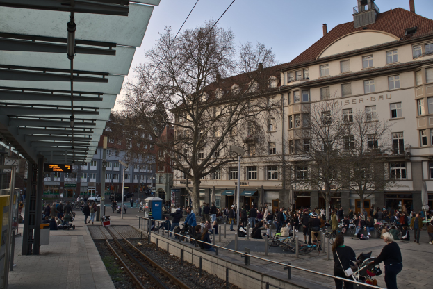Eine belebte Stadtstraße mit einem Bahnhof auf der linken Seite, umgeben von Gebäuden, Bäumen und Laternen, mit Menschen, die gehen, auf Bänken sitzen und in der Nähe der Station unter einem sichtbaren Himmel stehen.
