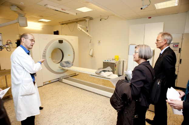Arzt in weißer Uniform erklärt eine CT-Scan-Maschine zwei Menschen in schwarzen Mänteln.