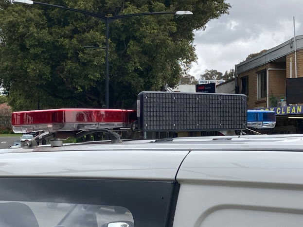 Ein Polizeiwagen mit eingeschaltetem Licht steht an der Seite einer Straße umgeben von Gebäuden, Bäumen, Pfählen und Texttafeln, mit einer bewölkten Himmel im Hintergrund, wahrscheinlich nach einem tödlichen Schulschießen in Brisbane.