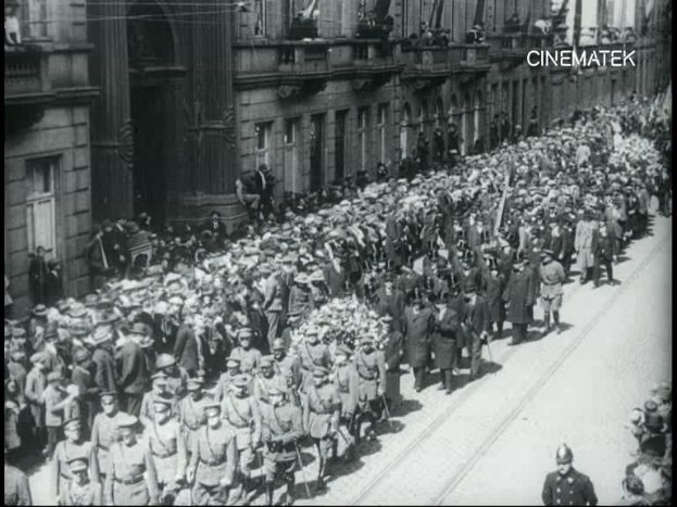Schwarzes und weißes Foto einer großen Menge Menschen, die vor einem Gebäude die Straße entlanggehen, einige halten Gewehre in den Händen, während es scheint, als würde ein Umzug stattfinden.