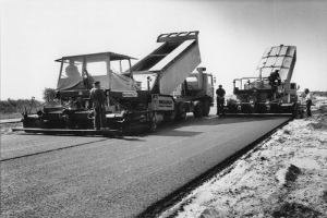 Schwarz-weiß-Foto einer Straße im Bau mit Arbeitern und Fahrzeugen, umgeben von Bäumen unter einem klaren Himmel.