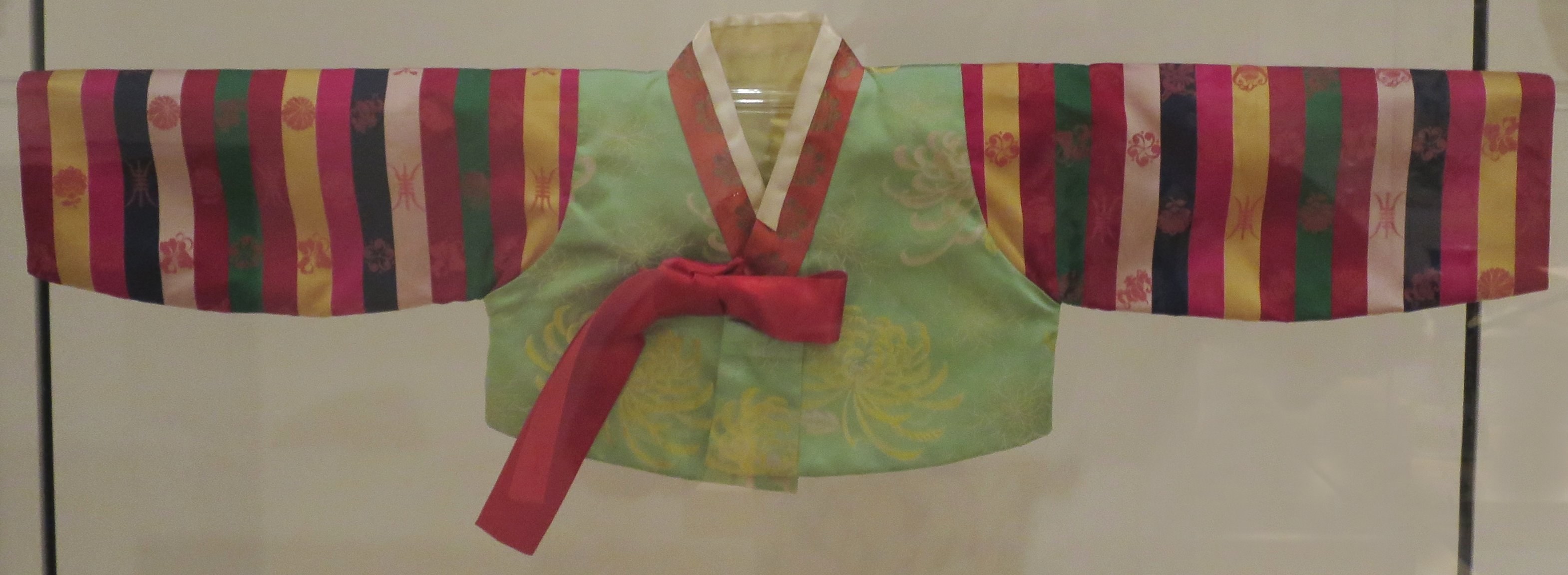Ein farbenfroher traditioneller koreanischer Hanbok mit kunstvollen Mustern in einer Glasvitrine auf einem wandmontierten Bügel.