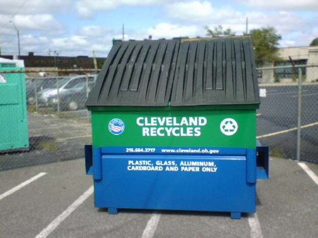 Ein Müllcontainer mit Logos und Text darauf steht auf dem Boden vor einem Zaun, mit parkenden Autos, Häusern und Bäumen im Hintergrund unter einem Himmel.
