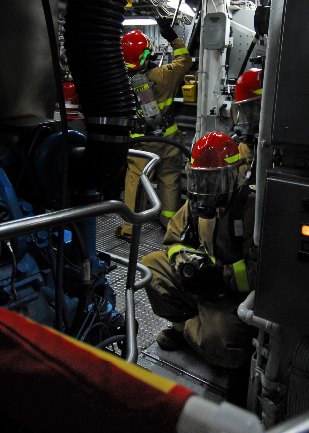 Feuerwehrleute in Helmen und Schutzausrüstung arbeiten an einem Löschfahrzeug mit Rohren und einer Lampe im Hintergrund.