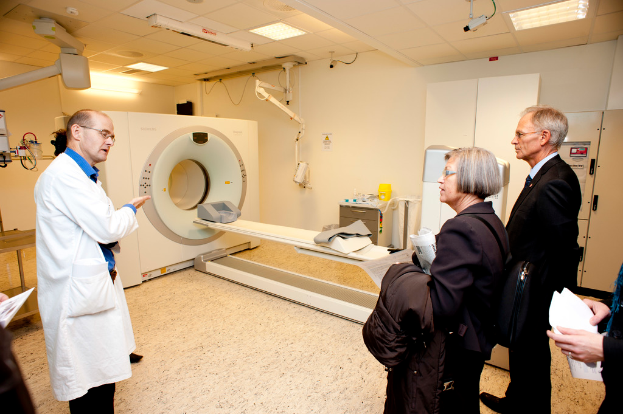 Ein Arzt in Weiß erklärt einem CT-Scan-Gerät zwei Personen in dunklen Anzügen.