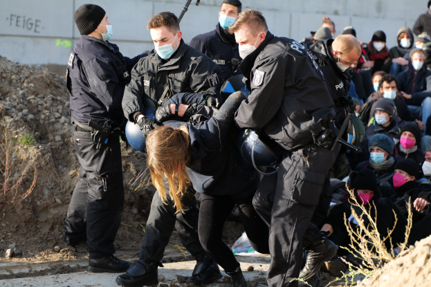 Gruppe von maskierten und mit Mützen bekleideten Personen, einige mit Waffen, die eine Frau umgeben, die von der Polizei eskortiert wird, mit Pflanzen, Steinen und einer Wand mit Text im Hintergrund.