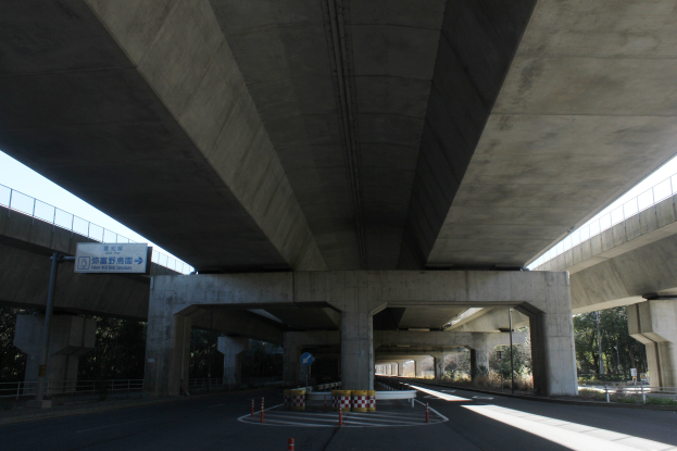 Autobahn unter einer Brücke mit Absperrkegeln, Barrierepfosten und Texttafeln entlang der Straße, flankiert von Geländern, mit Bäumen und einem klaren blauen Himmel im Hintergrund.