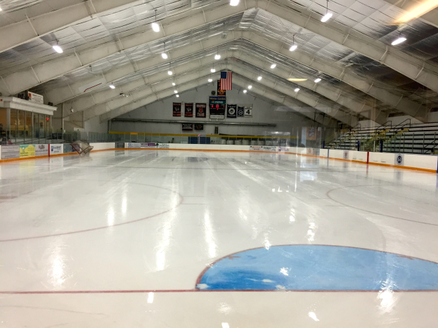 Große Indoor-Eisbahn mit Deckenbeleuchtung, Werbetafeln, einer Rückwand, Treppen mit Geländern und einigen Menschen, was auf eine Sportanlage hinweist, die likely für Hockey verwendet wird.