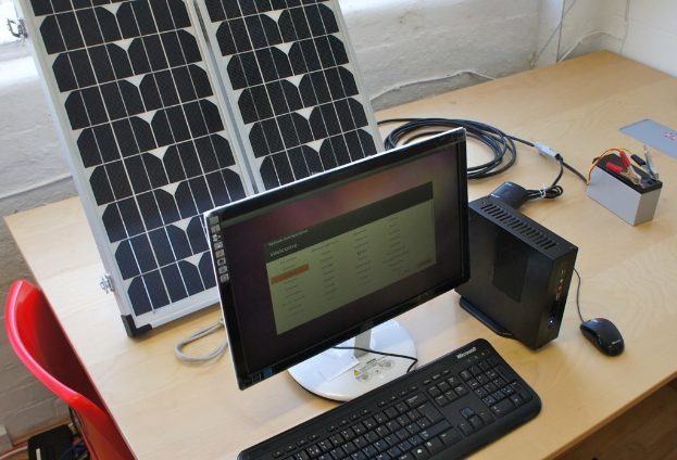 Ein Computersystem mit Tastatur, Maus und CPU auf einem Tisch, neben einer Solaranlage, Batterie, Chips und Kabeln, mit einem roten Stuhl und einer weißen Wand im Hintergrund.