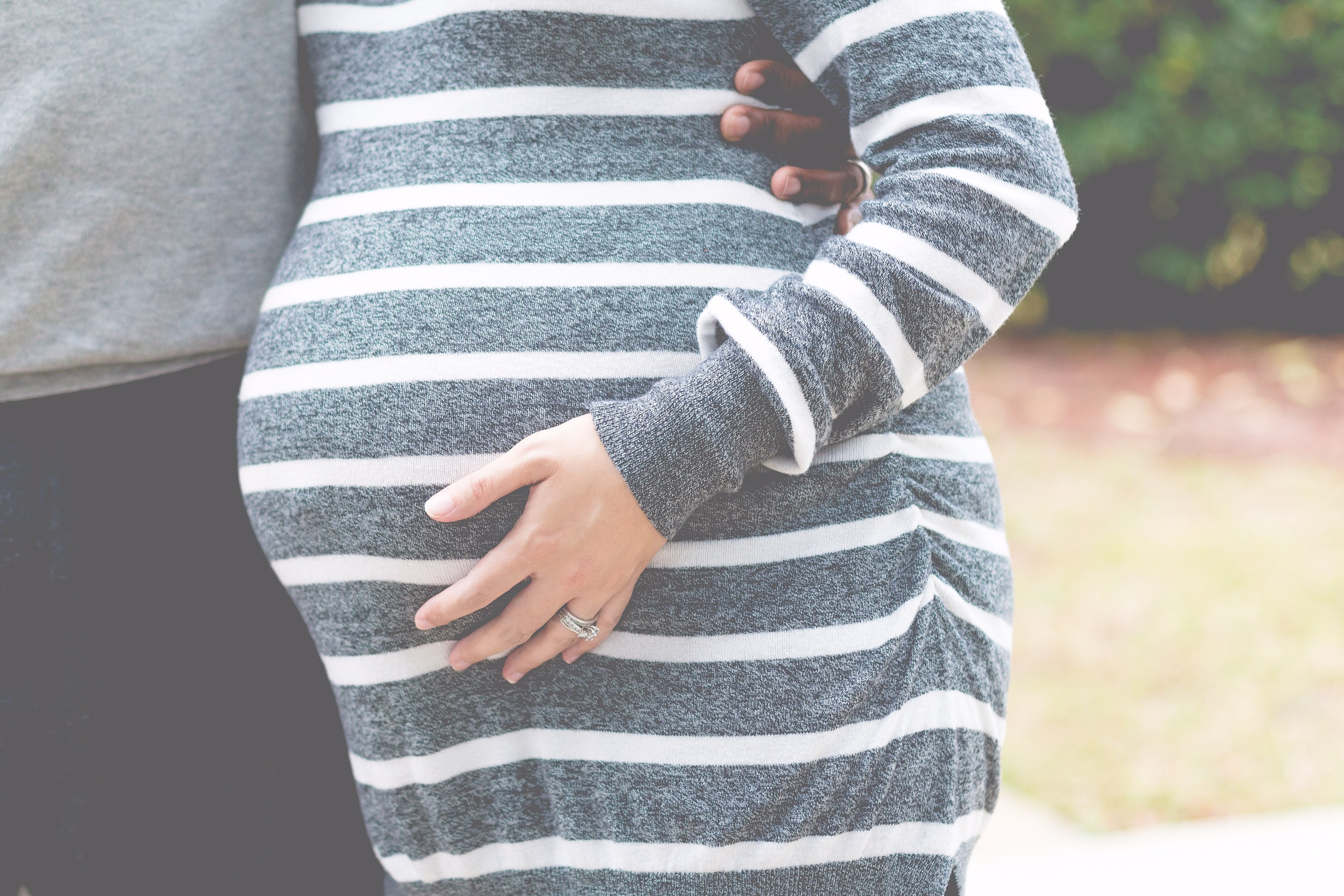 Schwangere Paare stehen nebeneinander mit den Händen auf ihrem Bauch, tragen Schwangerschaftskleidung, in einer holzigen Outdoor-Umgebung.