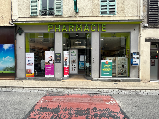 Eine Apotheke mit Glaswänden und -türen an einer Straßenecke, mit Plakaten an den Wänden und einer Straße im Vordergrund.