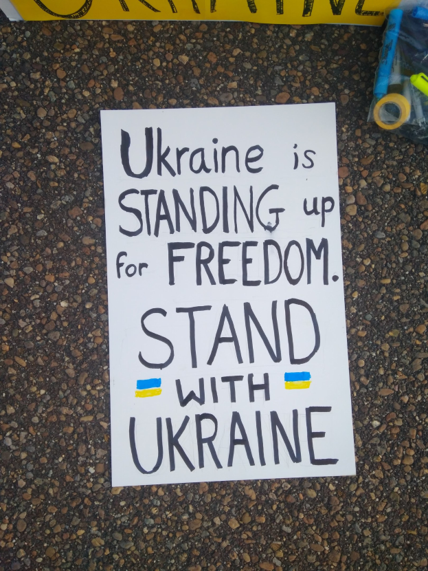 Ein weißer Papierbogen mit den Worten "Ukraine steht für Freiheit, steht mit Ukraine" liegt auf dem Boden vor einer gelben Fahne, rechts davon eine Plastikabdeckung mit verschiedenen Gegenständen.