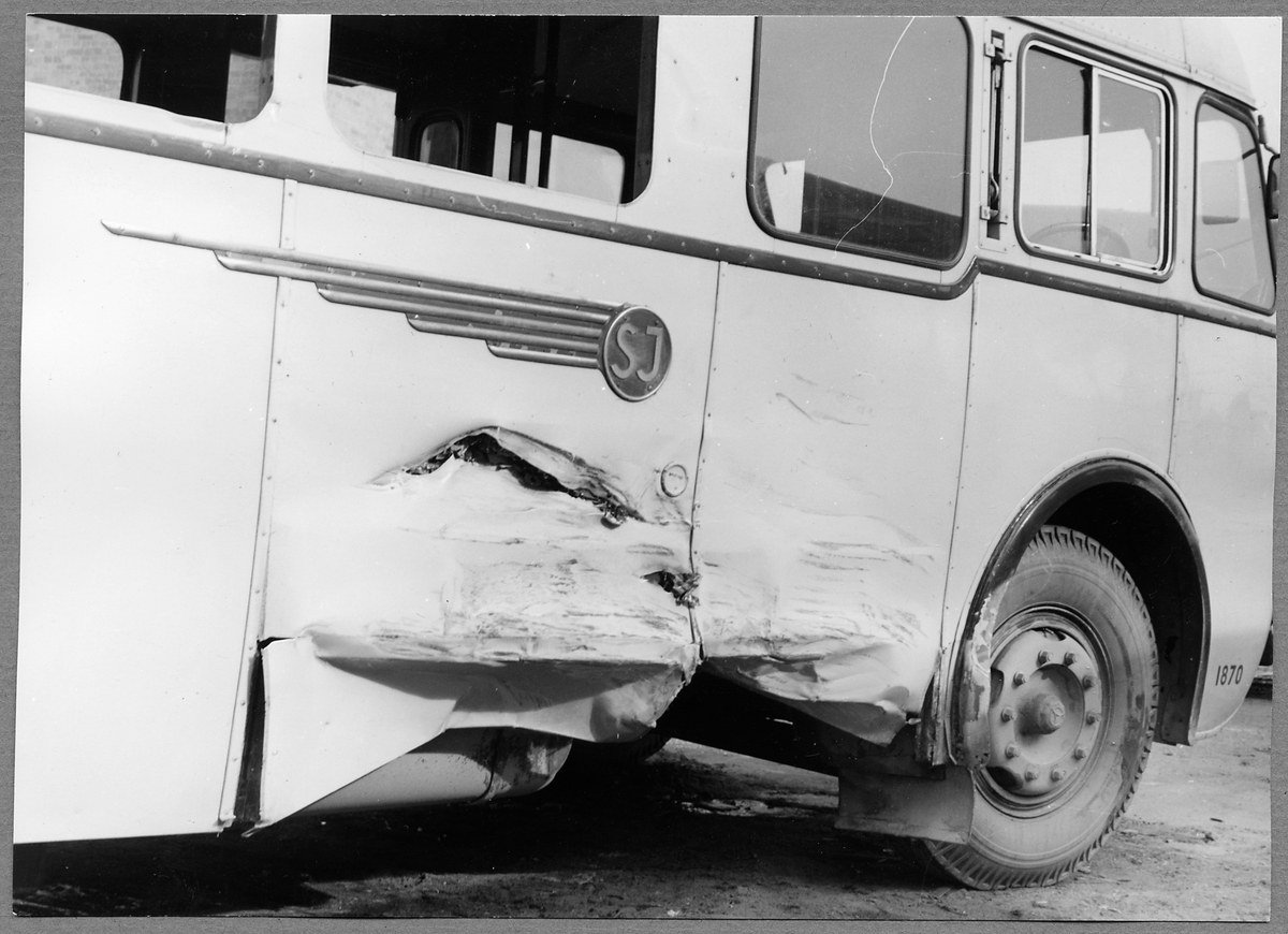 Ein Schwarz-Weiß-Foto eines beschädigten Busses mit eingedellter Front, zersplitterten Fenstern und geöffneter Fahrertür auf der Straße.