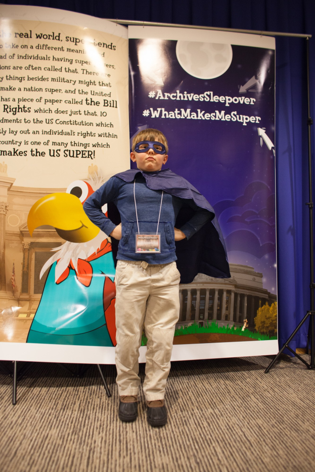 Ein junger Junge in einem Superheldenkostüm steht vor einem Banner mit Text und einem Vogelbild, mit einem Vorhang dahinter.