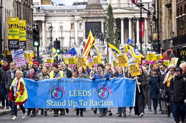 Eine Gruppe von Menschen marschiert mit einer 'Steh Auf gegen Rassismus'-Schlagzeile die Straße entlang, mit Gebäuden, Bäumen, Laternen, Verkehrszeichen, einer Statue und einem bewölktem Himmel im Hintergrund.