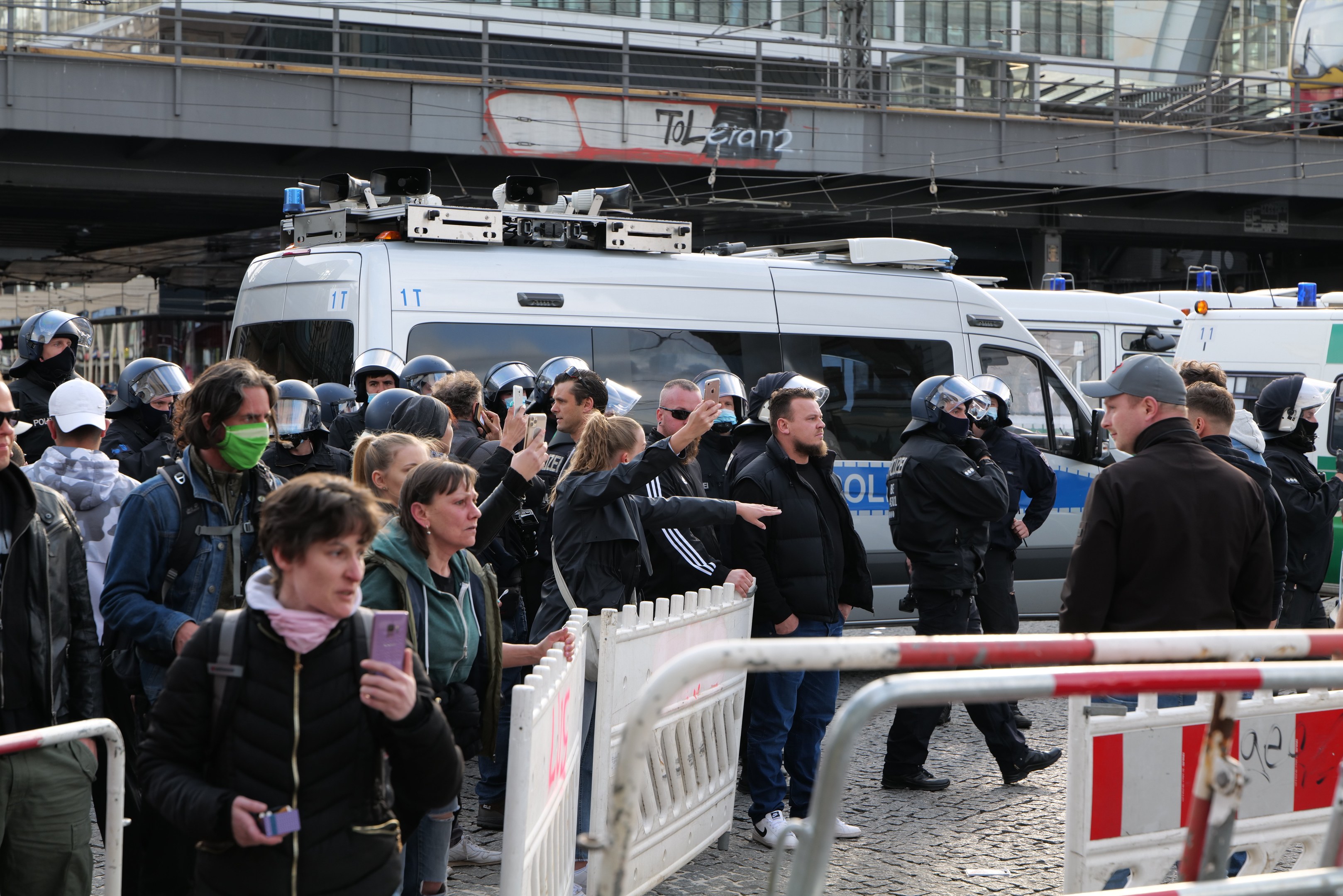 Eine Gruppe von Menschen steht vor Polizeifahrzeugen, einige tragen Helme und halten Telefone, mit Barrieren im Vordergrund, einer Brücke und Gebäuden im Hintergrund, während einer Protestauflösung in Berlin, Deutschland.