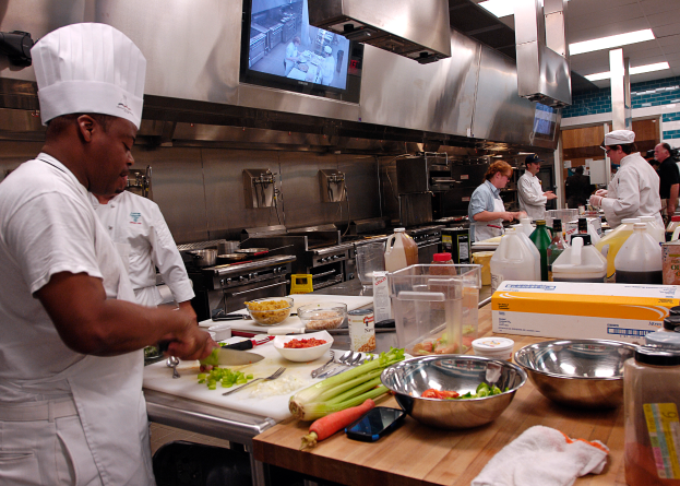 Eine Gruppe von Köchen in einer Küche, die Essen auf einem Schneidbrett zubereiten, mit einem Koch im Vordergrund in einer weißen Schürze und Mütze, der ein Messer hält, und umgeben von verschiedenen Utensilien, Schüsseln, Gemüse, einem Handy, Flaschen und anderen Gegenständen, mit einem Fernseher und Deckenleuchten im Hintergrund.