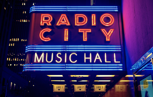 Radio City Music Hall in New York City, hell erleuchtet in der Nacht mit hohen Gebäuden drumherum und einem "Radio City"-Schild auf der rechten Seite.