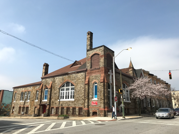Ein altes Backsteingebäude, das als erste Baptistenkirche der Stadt identifiziert wurde, wird an einer Straßenecke renoviert, mit sichtbaren Fenstern, Laternen, Verkehrszeichen, Schildern, Bäumen, Fahrzeugen und einem bewölkten Himmel im Hintergrund.