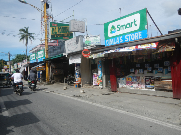 Eine belebte Straße mit Motorrädern, Autos, Gebäuden, Straßenschildern, Strommasten, Kabeln, Straßenlaternen, Bäumen und einem bewölkten Himmel.