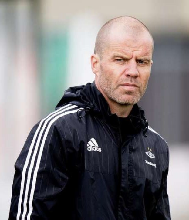 Ein kahlköpfiger Trainer in einer blauen Jacke mit weißen Streifen und einem weißen Kragen steht auf einem Fußballfeld und hat die Hände verschränkt und einen ernsten Gesichtsausdruck.