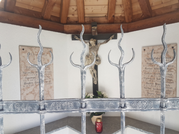Ein Metallzaun mit einer Statue von Jesus darauf, umgeben von einem Blumenkübel und anderen Gegenständen auf dem Boden, mit Tafeln mit Text und Zahlen an der Wand dahinter und einem hölzernen Dach oben am Holocaust-Mahnmal in Stuttgart, Deutschland.