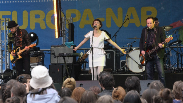 Vier Musiker auf der Bühne: Eine Frau in einem weißen Kleid singt in der Mitte, flankiert von zwei Männern mit Gitarren, mit einer Menge unten und Lautsprechern im Hintergrund an der Wand.