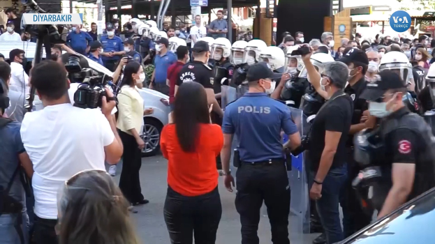 Eine Gruppe von Menschen vor einer Reihe von Polizisten in Helmen, einige mit Kameras, mit Fahrzeugen, Gebäuden und Schildern mit der Aufschrift "Türkische Polizei gerät mit Demonstranten in Ankara aneinander".