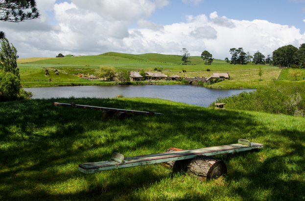 Stämme mit Holzplanken darauf, eine grüne Landschaft mit einem Teich in der Mitte, Häuser, Bäume und Hügel im Hintergrund unter einem Himmel.