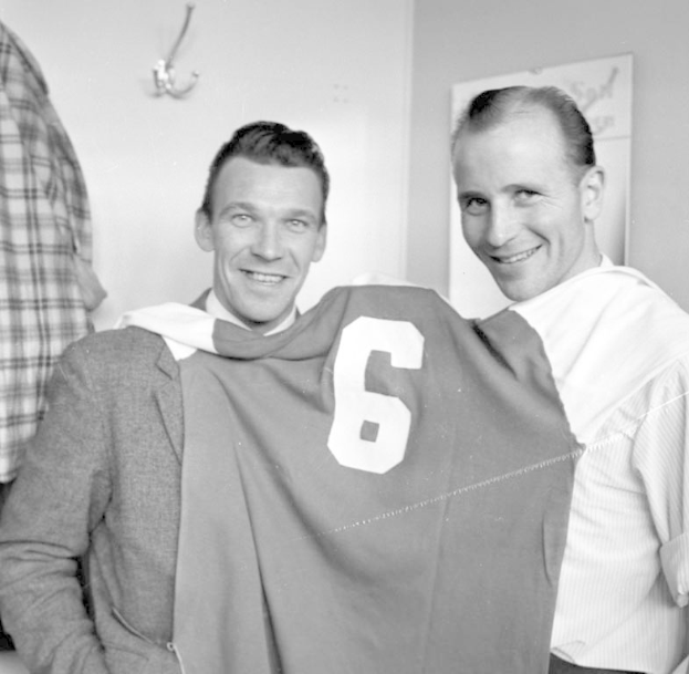 Zwei Männer in Schwarz-Weiß, die strahlend eine Fußballtrikot hochhalten, mit einer Tafel und einem Bügel an der Wand dahinter.