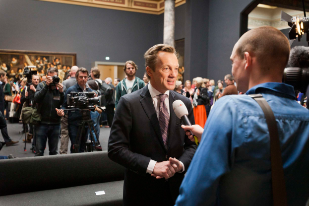 Ein Mann im Anzug und Krawatte spricht mit einem Reporter vor einer Menge, einige halten Kameras, mit einem Sofa im Vordergrund und einer Wand mit einem Foto Rahmen, Lampen und einer Säule im Hintergrund, hält ein Mikrofon.