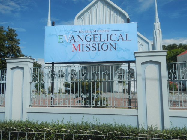 Kirche mit Metallzaun, Schild "Evangelische Mission", umgeben von Bäumen, Haus mit Dach und Pflanzen unter einem bewölkten Himmel.