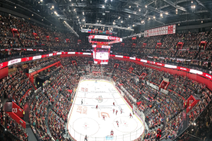 Eine große, hell erleuchtete Arena voller Zuschauer, die ein Hockey-Spiel sehen, mit einem zentralen Bildschirm über der Eisfläche und einer elektrisierenden Atmosphäre.