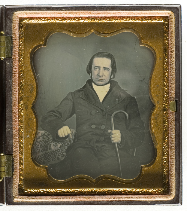 Ein altes Daguerreotyp eines Mannes in einem Anzug, der in einem Stuhl sitzt und einen Stock hält, mit einem ernsten Gesichtsausdruck und konzentriertem Blick.