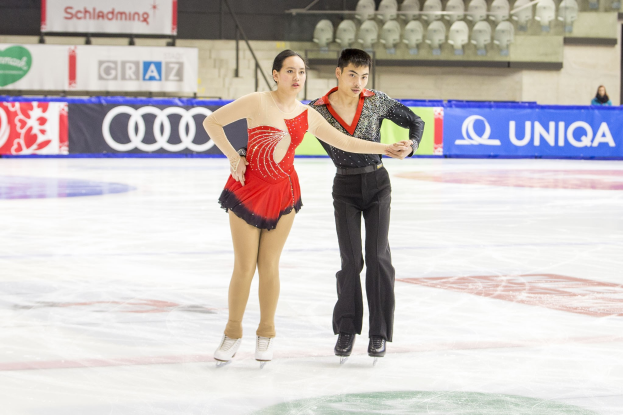 Zwei Eiskunstläufer, ein Mann und eine Frau, gleiten Hand in Hand über eine Eisbahn mit einigen Zuschauern und Event-Elementen im Hintergrund.