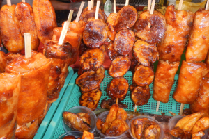Marktstand mit Spießen von Hähnchenflügeln, verschiedenen Lebensmitteln in Plastikbehältern, mit Menschen und Flaschen im Hintergrund.