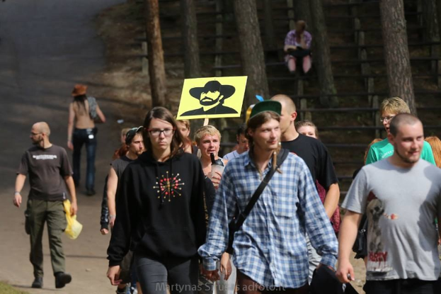 Eine Gruppe von Menschen, die einen Waldweg entlanggehen, einige tragen Taschen und einer hält ein Schild mit einem Personenbild, mit Stufen und Geländern im Hintergrund.