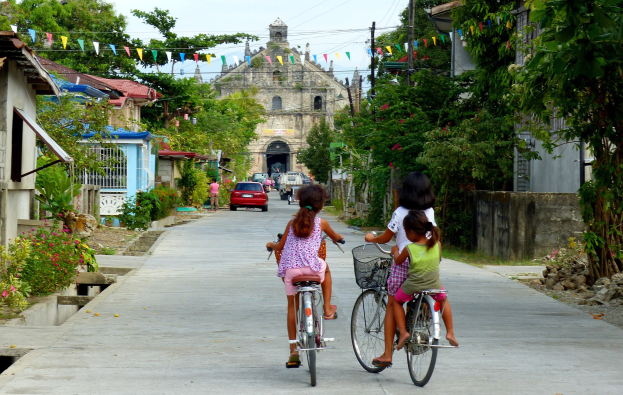 Drei Kinder fahren Fahrräder auf einer von Bäumen gesäumten Straße mit Gebäuden auf beiden Seiten, ein Auto und ein Gebäude sind im Hintergrund und der Himmel darüber sichtbar.