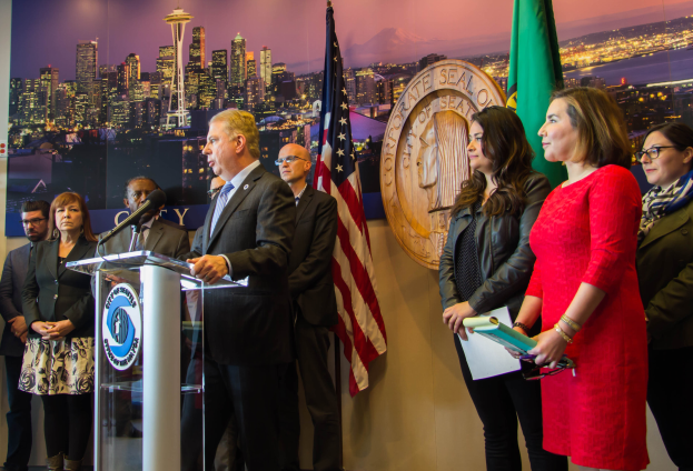 Eine Gruppe von Menschen um ein Rednerpult vor einer Wand mit Flaggen und einer Tafel, auf der 'Seattle Mayor Bill de Blasio und die Stadt Seattle City Council' steht; ein Mann im Anzug steht am Rednerpult und spricht in ein Mikrofon, und eine Frau rechts von ihm hält Gegenstände in den Händen.