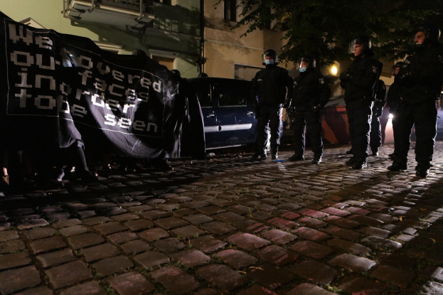 Eine Gruppe von Polizisten in Helmen, die ein Banner auf einer Kopfsteinpflasterstraße bei Nacht halten, mit Fahrzeugen, Gebäuden, Bäumen und Straßenbeleuchtung im Hintergrund.