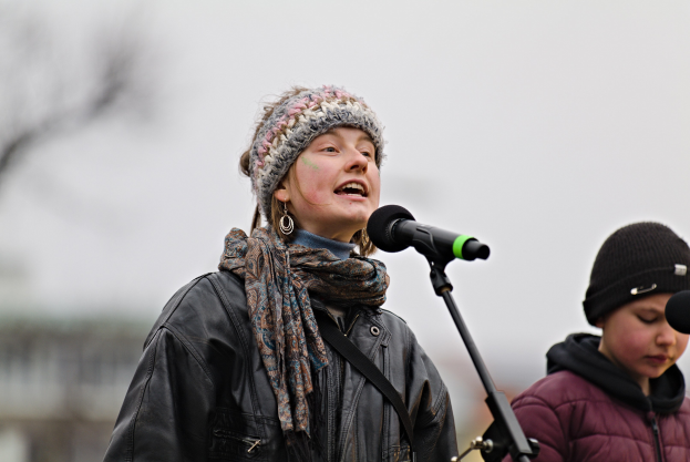 Eine Frau in schwarzer Jacke, Schal und Mütze singt in ein Mikrofon auf der Bühne, während eine andere Person in einer maronenfarbenen Jacke und schwarzer Mütze neben ihr steht und ein unscharfer Hintergrund die Menge bildet.