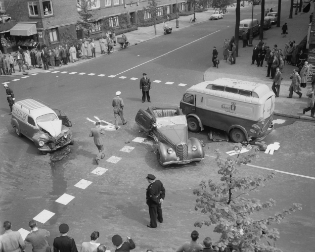 Schwarz-weißes Bild einer Autounfall-Szene an einer Straßenkreuzung mit mehreren Fahrzeugen, darunter ein Lieferwagen, und einer Gruppe von Schaulustigen; Gebäude, Bäume, Strommasten und Texttafeln sind im Hintergrund zu sehen.