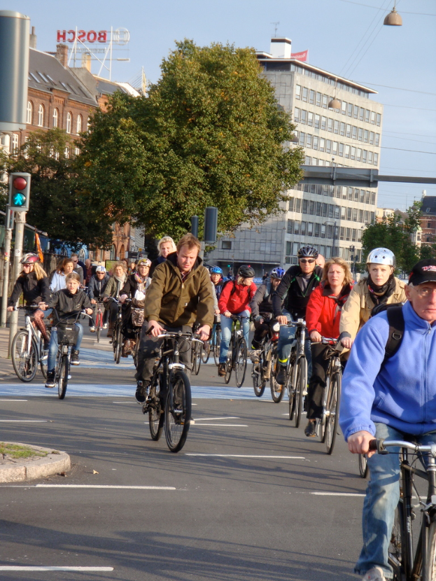 Menschen fahren Rad auf einer Straße mit Pfählen, Bäumen, Gebäuden und dem Himmel im Hintergrund.