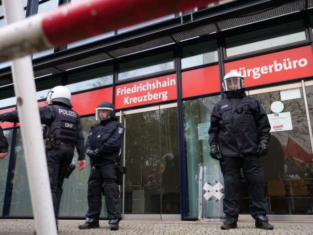 Eine Gruppe von Polizisten in Helmen und Handschuhen steht vor einem gläsernen Gebäude, mit einem Pfahl links und Schildern im Hintergrund, auf denen "Friedrichshain Kreuzberg" steht, und Papieren an den Glaswänden.
