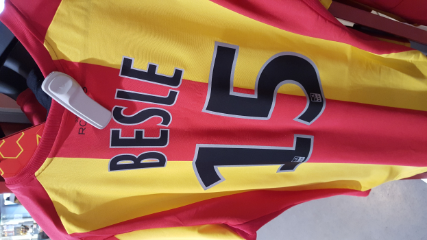 Ein roter und gelber Fußballtrikot mit Text und Nummern hängt an einem Ständer in einem Geschäft, mit anderen Gegenständen und dem Boden im Hintergrund.