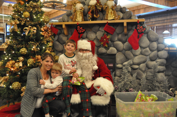 Eine Frau, Kinder und ein als Santa Claus verkleideter Mann mit Brille und Handschuhen stehen neben einem geschmückten Weihnachtsbaum, einer Wand mit Felsen, Spielzeug, Dekorationen und einem Korb mit Gegenständen, mit Lichtern im Hintergrund.