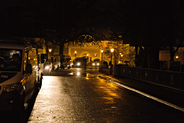 Eine nächtliche Stadtstraße mit parkenden Autos, beleuchtet von Straßenlaternen, gesäumt von Bäumen und einem Gebäude im Hintergrund.