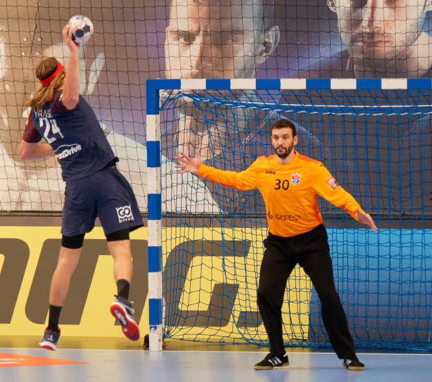 Zwei Männer spielen Handball vor einem Tor, mit einem Netz und einer Fahne mit Text und Bildern von Menschen im Hintergrund.
