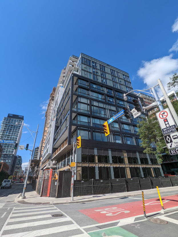 Eine belebte Stadtstraße mit einem hohen Wohn-/Geschäftshaus zum Verkauf an der 541 East 57th Street, Einheit 541, Fahrzeugen auf der Straße, Verkehrszeichen, Schilder, Bäumen, einem Zaun und einem bewölkten Himmel.