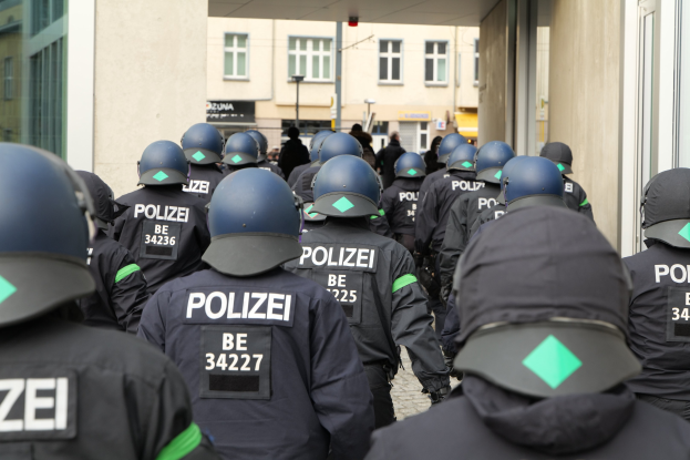 Eine Gruppe von Polizisten in Uniform und Helmen vor einem Gebäude stehend, mit einigen Menschen und Gebäuden im Hintergrund.